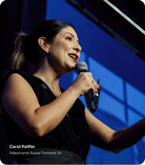 Carol Paiffer palestrando no palco da edição do Asaas Connect de 2024. Ela está de preto, segurando um microfone e sorrindo.