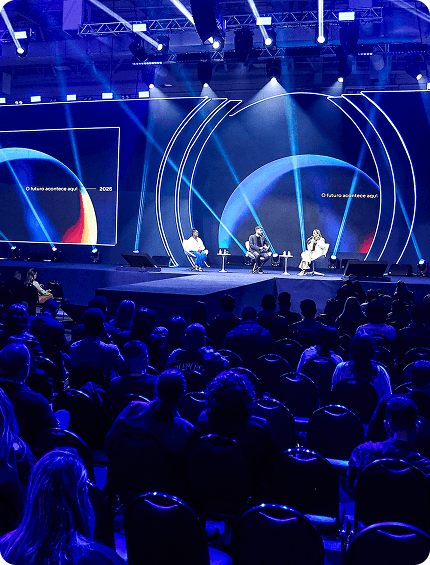 Palestrantes no Palco Connect. O palco é cheio de luzes azuis e possui um grande telão.